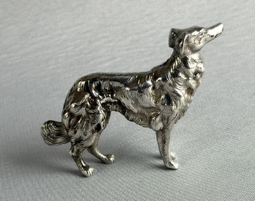 fig 1. A small cast silver figure of a wolfhound standing in profile on a plain light grey surface.