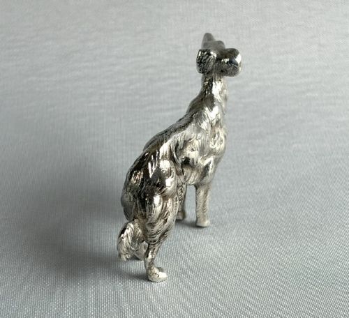 A small cast silver figure of a wolfhound standing in profile on a plain light grey surface. View IV