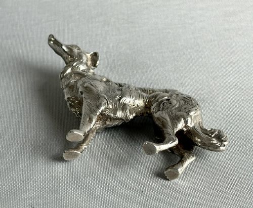 A small cast silver figure of a wolfhound standing in profile on a plain light grey surface. View V
