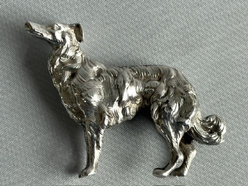 A small cast silver figure of a wolfhound standing in profile on a plain light grey surface. View VI