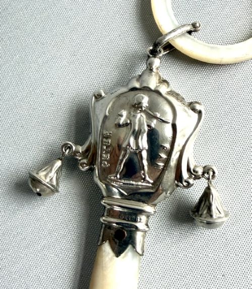fig 1. An antique sterling silver baby rattle featuring an embossed Peter Pan motif, two attached side bells, a mother-of-pearl handle, and a top mother-of-pearl teething ring.