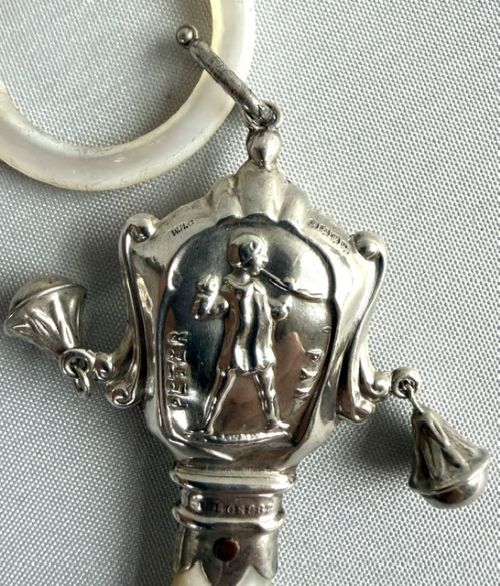 An antique sterling silver baby rattle featuring an embossed Peter Pan motif, two attached side bells, a mother-of-pearl handle, and a top mother-of-pearl teething ring. View IV