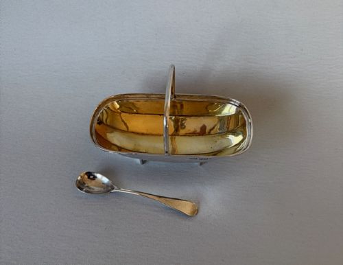 fig 1. An Edwardian sterling silver trug-shaped condiment dish featuring a gilded interior, central arched handle, four small feet, and an accompanying silver spoon.