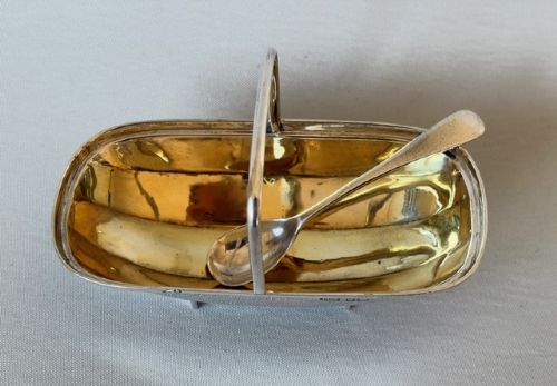 An Edwardian sterling silver trug-shaped condiment dish featuring a gilded interior, central arched handle, four small feet, and an accompanying silver spoon. View IV