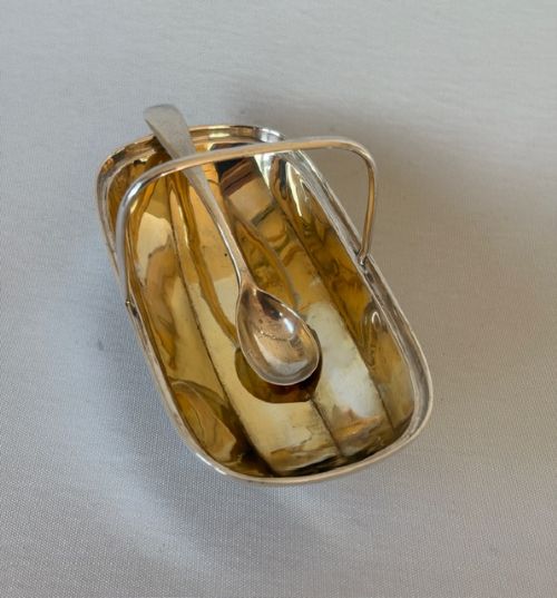 An Edwardian sterling silver trug-shaped condiment dish featuring a gilded interior, central arched handle, four small feet, and an accompanying silver spoon. View V