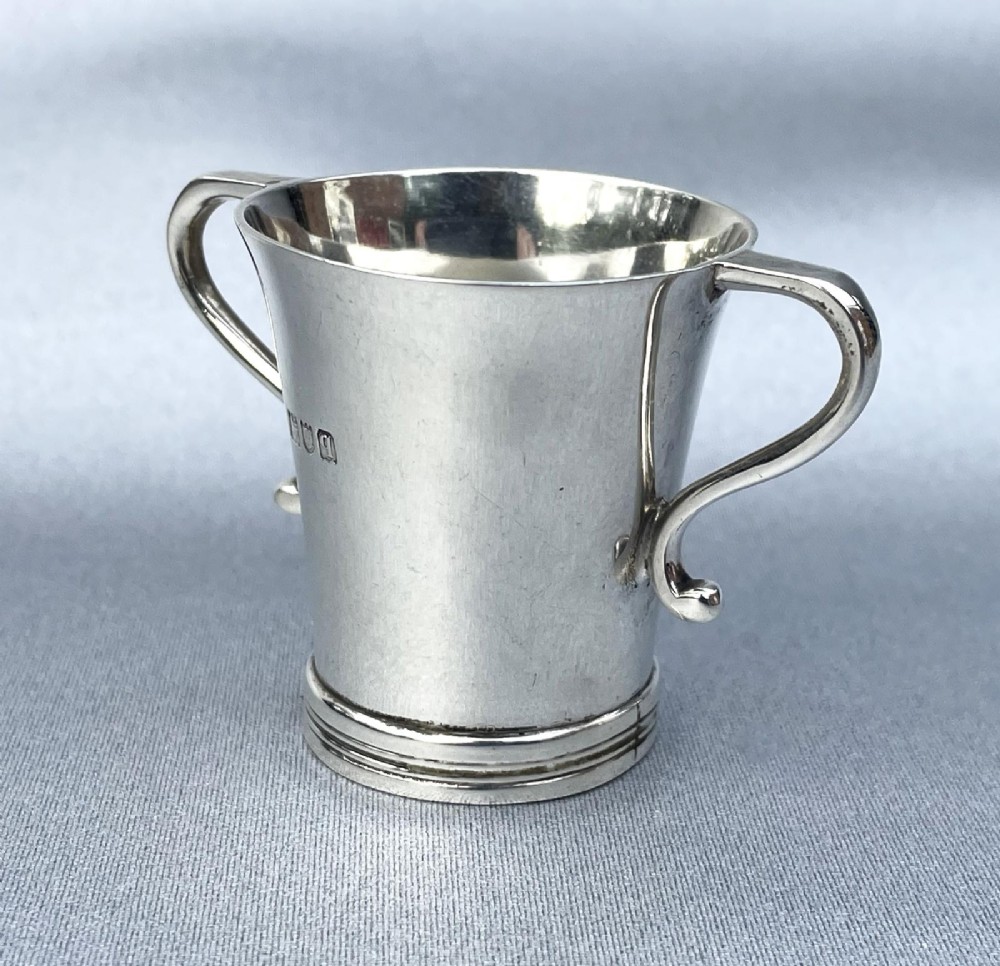 A small George V sterling silver tot cup measure featuring twin scroll handles and visible hallmarks on the exterior body.