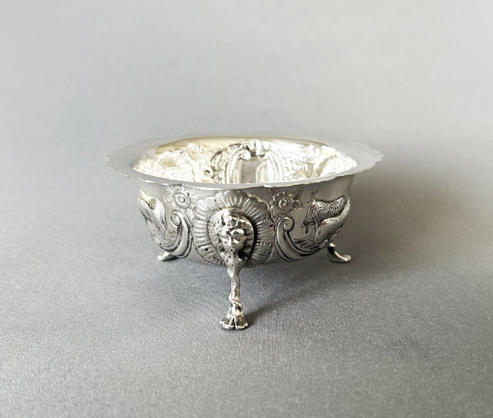 A circular silver bowl featuring embossed goat motifs, floral repousse decoration, a scalloped rim, and three sculptural mask feet.