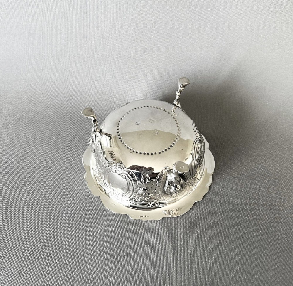 A circular silver bowl featuring embossed goat motifs, floral repousse decoration, a scalloped rim, and three sculptural mask feet. View III