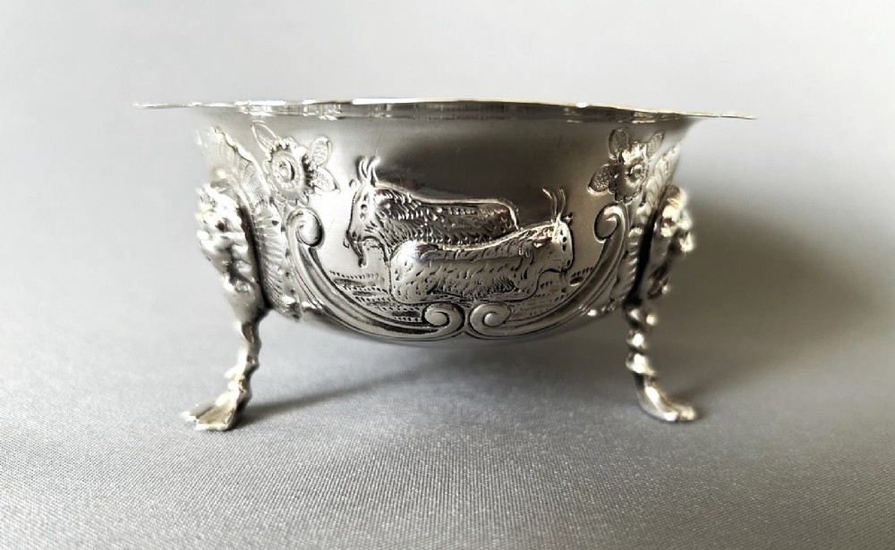 A circular silver bowl featuring embossed goat motifs, floral repousse decoration, a scalloped rim, and three sculptural mask feet. View IV