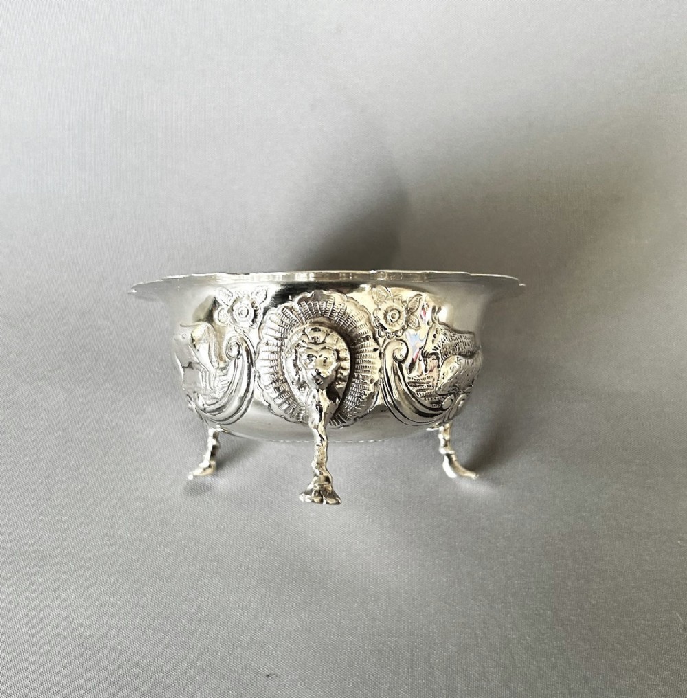 A circular silver bowl featuring embossed goat motifs, floral repousse decoration, a scalloped rim, and three sculptural mask feet. View V