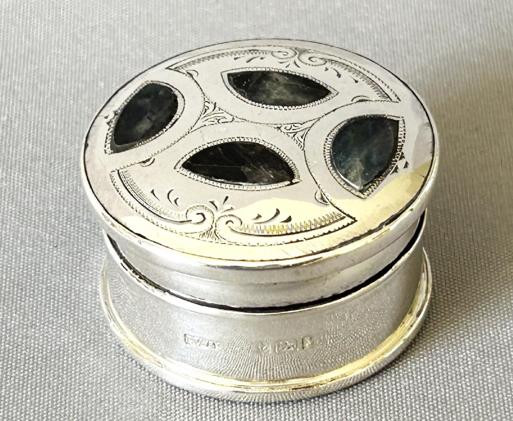 A circular sterling silver pillbox featuring engraved scrollwork on the lid surrounding four inlaid dark green agate stones.