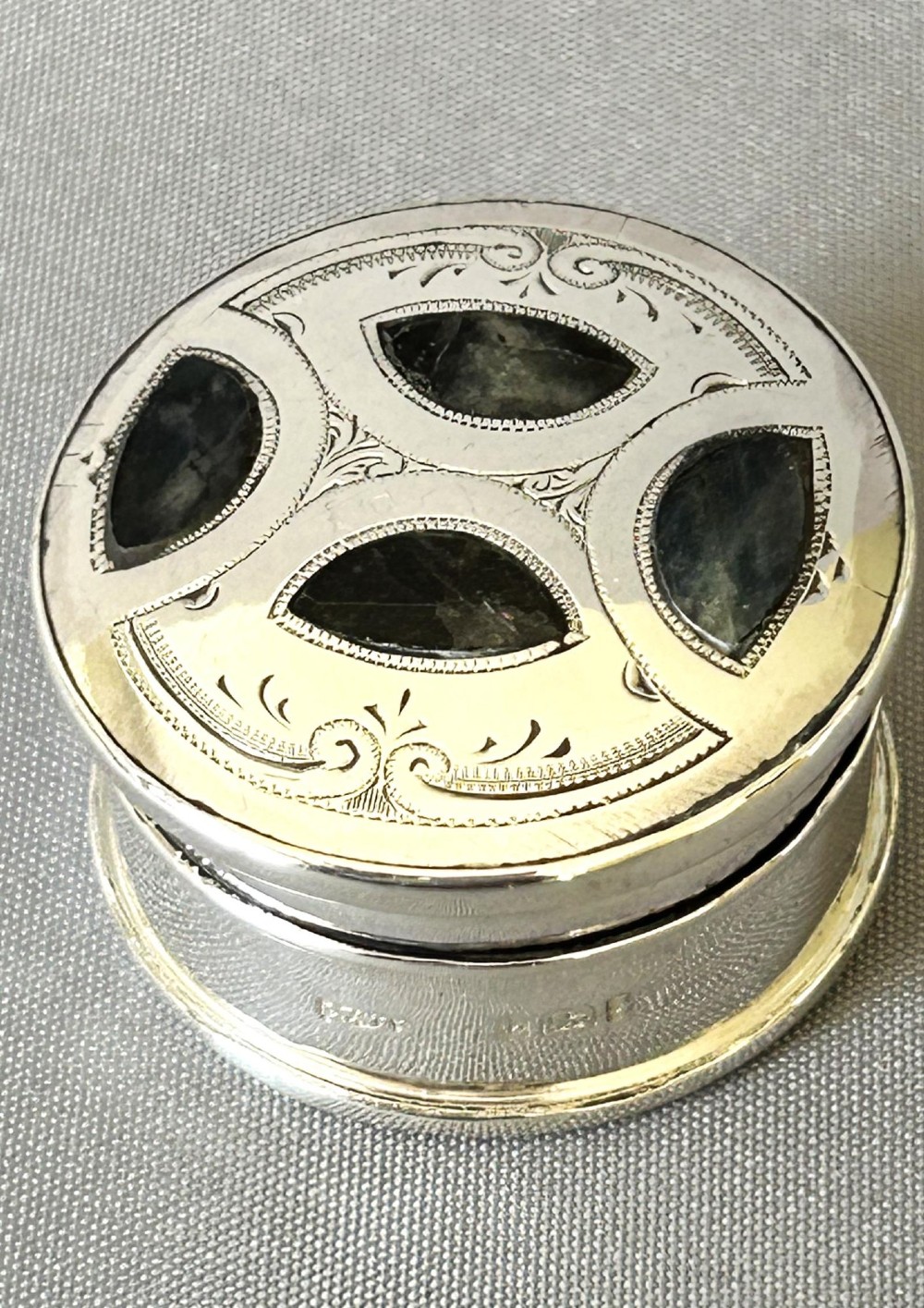 A circular sterling silver pillbox featuring engraved scrollwork on the lid surrounding four inlaid dark green agate stones. View III