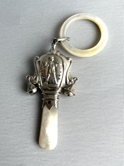 An antique sterling silver baby rattle featuring an embossed Peter Pan motif, two attached side bells, a mother-of-pearl handle, and a top mother-of-pearl teething ring. View V