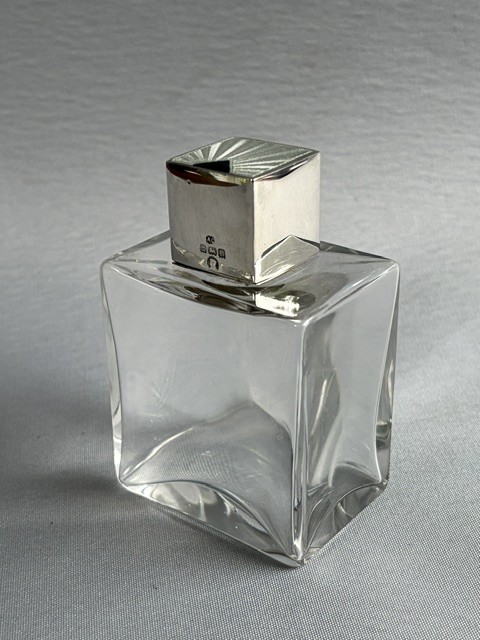 An Art Deco clear glass cologne bottle featuring a cubic silver cap with engine-turned sunburst detailing.