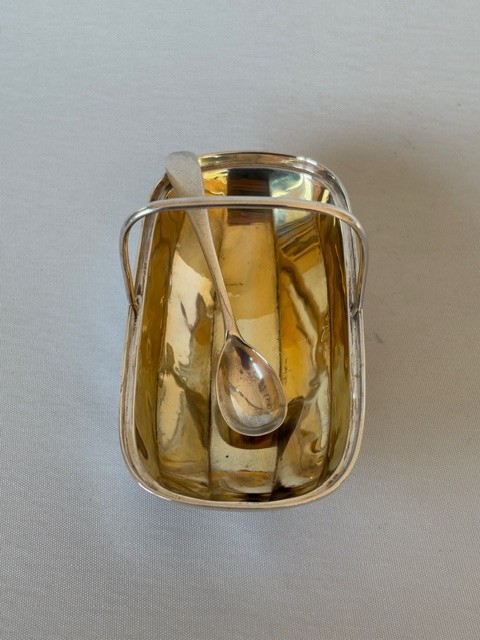 An Edwardian sterling silver trug-shaped condiment dish featuring a gilded interior, central arched handle, four small feet, and an accompanying silver spoon. View IX