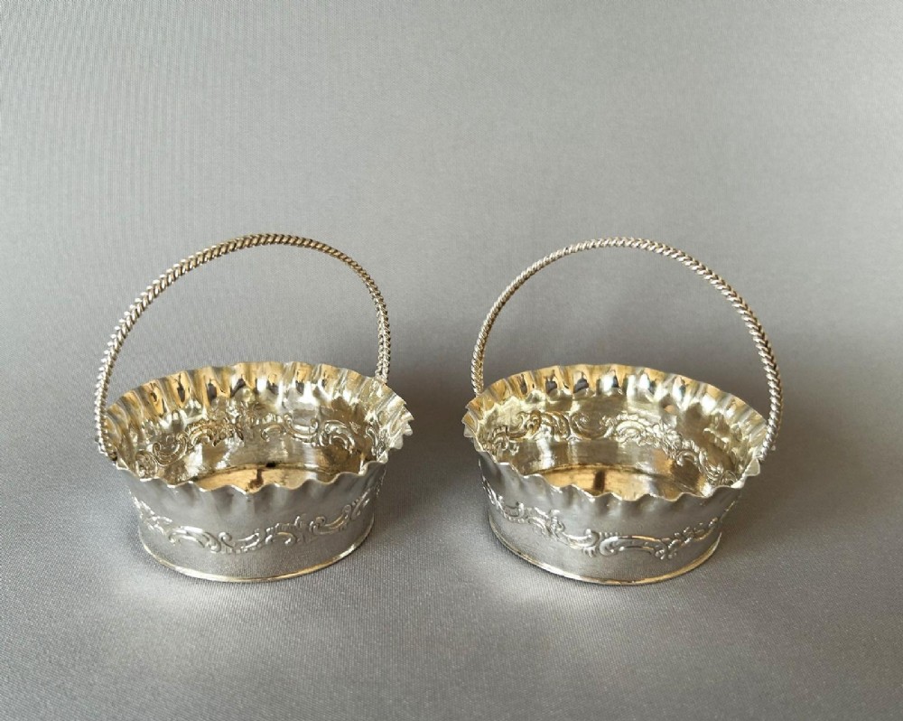 A pair of small silver bonbon baskets featuring crimped rims, embossed scrollwork motifs, and twisted overhead rope handles.