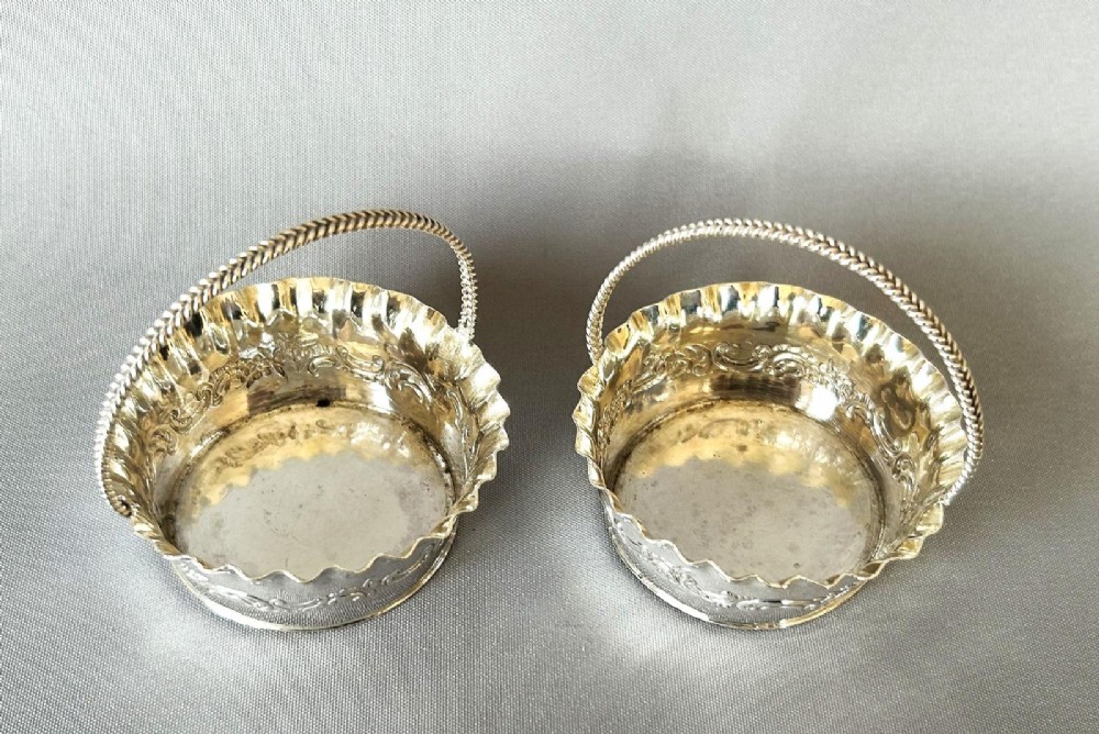 A pair of small silver bonbon baskets featuring crimped rims, embossed scrollwork motifs, and twisted overhead rope handles. View IV