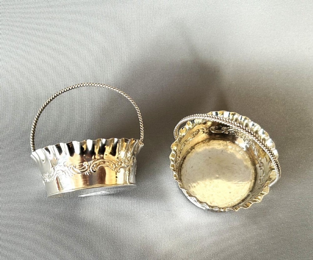 A pair of small silver bonbon baskets featuring crimped rims, embossed scrollwork motifs, and twisted overhead rope handles. View V