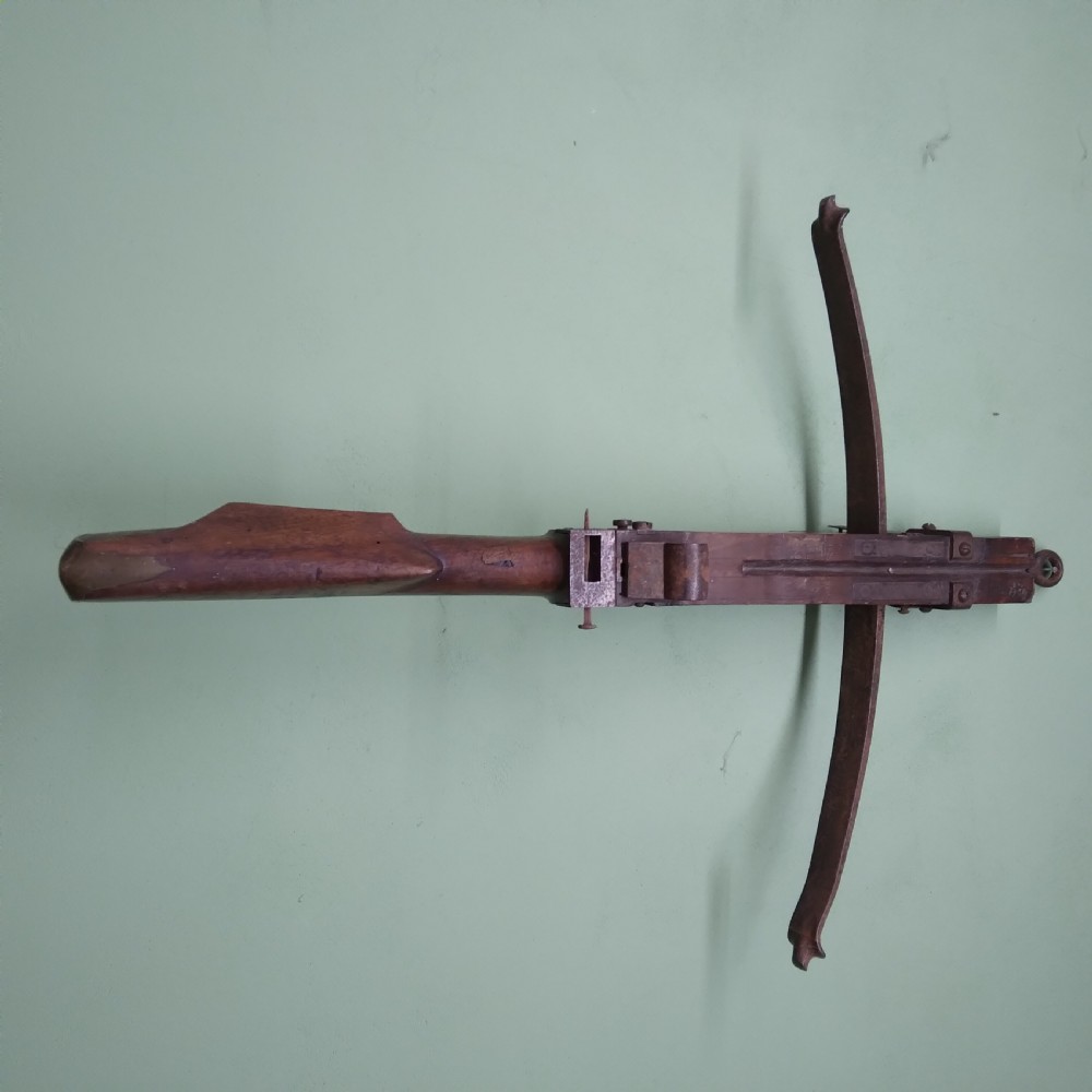 An antique wooden crossbow featuring a carved dark timber stock, a curved iron bow prod, and brass trigger embellishments. View V