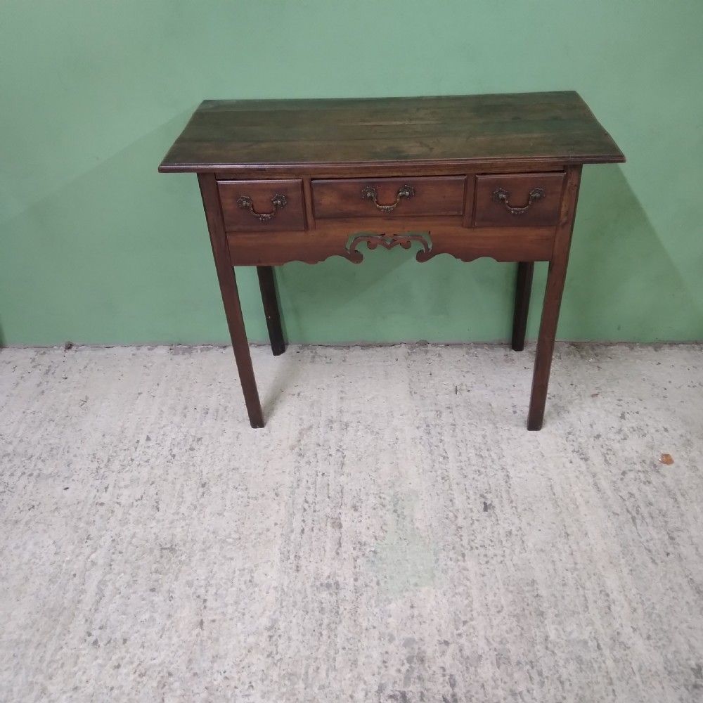 An antique fruitwood lowboy table featuring three drawers with brass drop handles, a shaped lower apron, and four square legs.