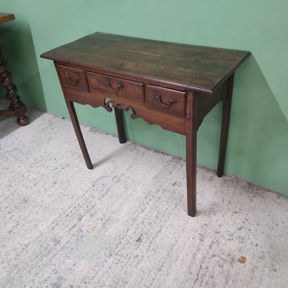 An antique fruitwood lowboy table featuring three drawers with brass drop handles, a shaped lower apron, and four square legs. View II