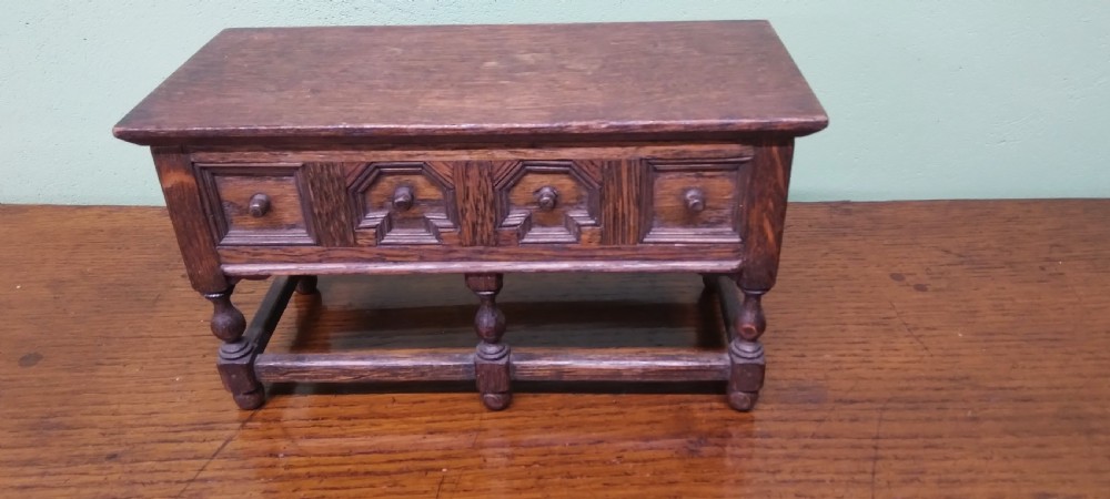 A small dark oak miniature dresser featuring four carved geometric panel fronts, small wooden knob handles, turned legs, and a bottom stretcher rail.