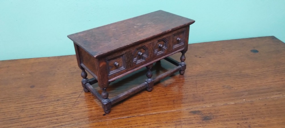 A small dark oak miniature dresser featuring four carved geometric panel fronts, small wooden knob handles, turned legs, and a bottom stretcher rail. View II