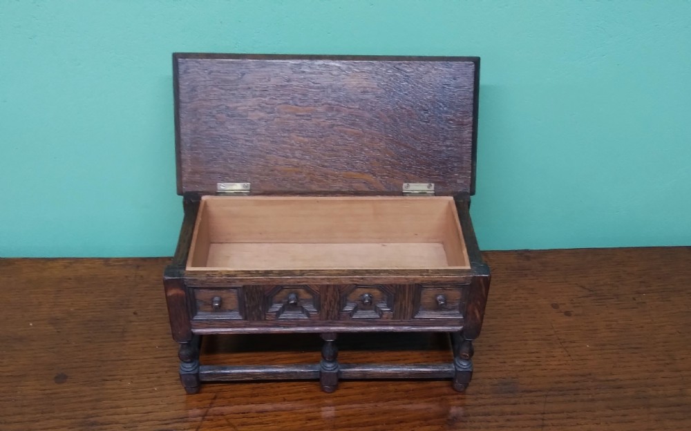 A small dark oak miniature dresser featuring four carved geometric panel fronts, small wooden knob handles, turned legs, and a bottom stretcher rail. View IV