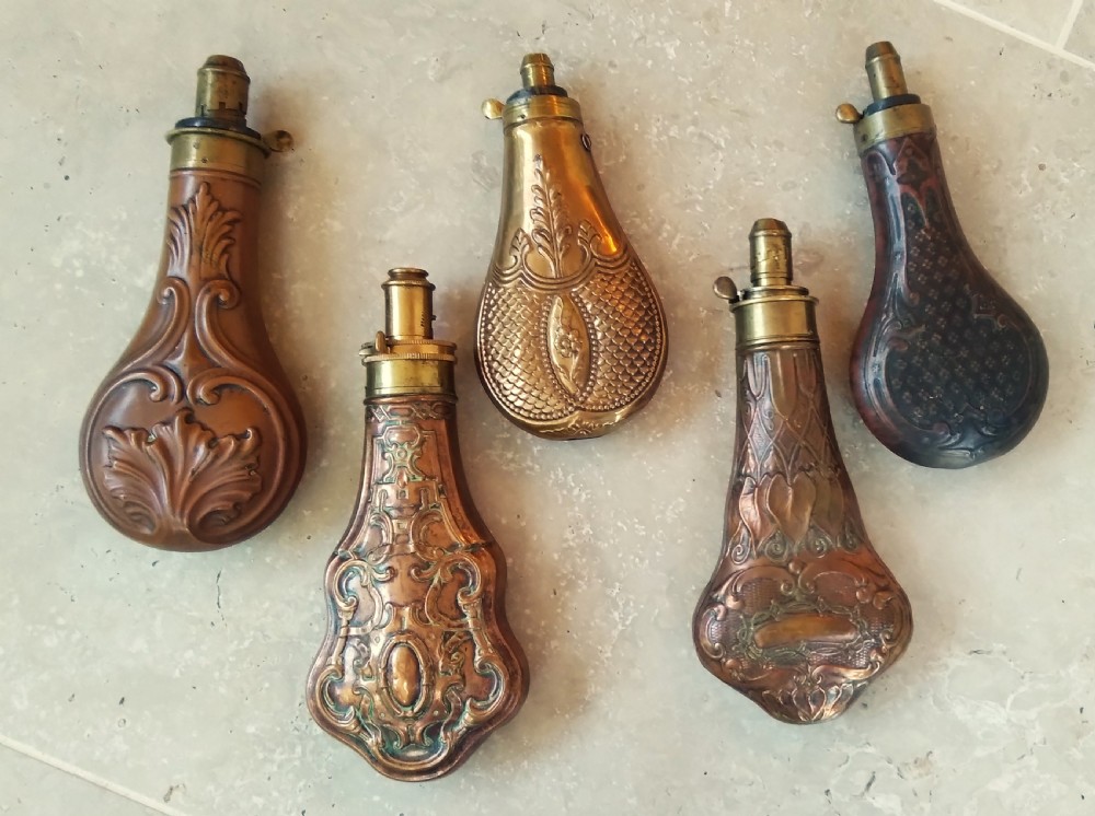 A collection of five 19th-century embossed brass and copper shot flasks featuring spring nozzles and intricate relief designs.