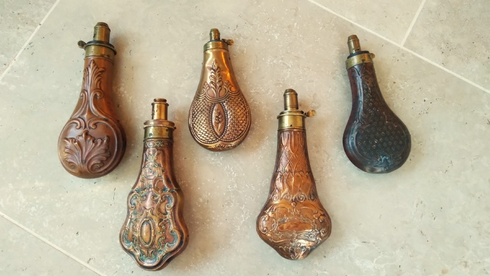 A collection of five 19th-century embossed brass and copper shot flasks featuring spring nozzles and intricate relief designs. View VII