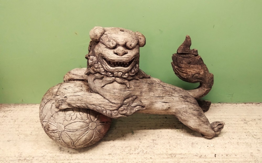A weathered hand-carved wooden Chinese guardian lion resting its front paw on a decorative brocade ball.