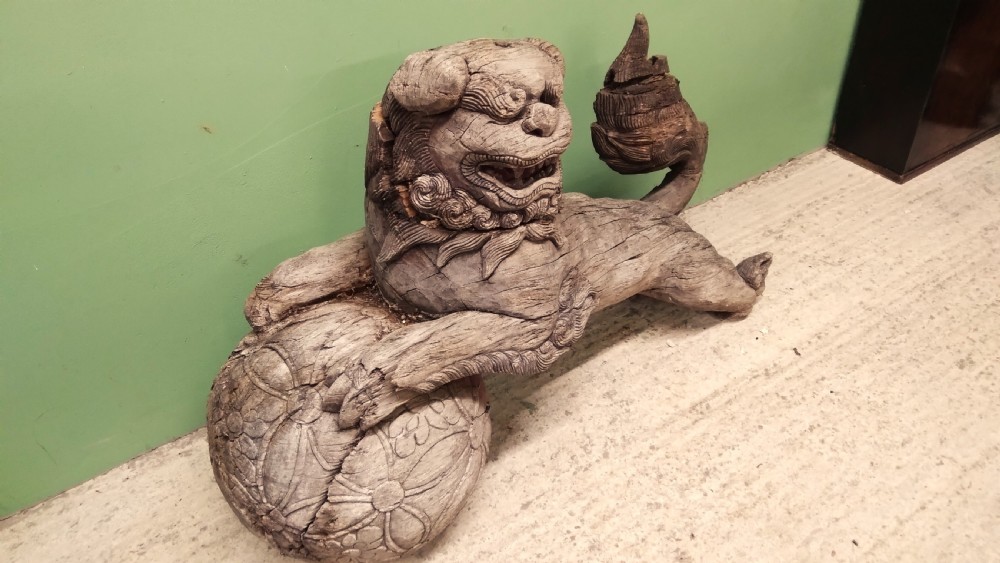 A weathered hand-carved wooden Chinese guardian lion resting its front paw on a decorative brocade ball. View II
