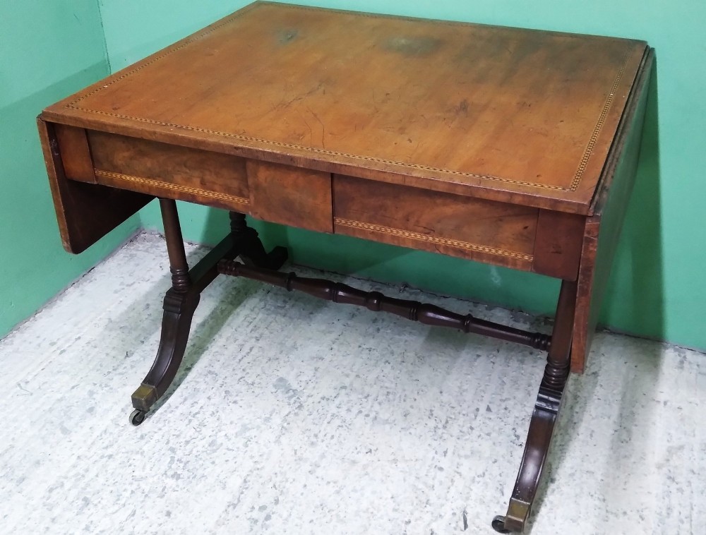 A George III mahogany drop-leaf sofa table featuring decorative border string inlay, two front drawers, a turned stretcher, and splayed legs fitted with brass castors.