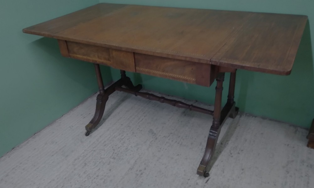 A George III mahogany drop-leaf sofa table featuring decorative border string inlay, two front drawers, a turned stretcher, and splayed legs fitted with brass castors. View II