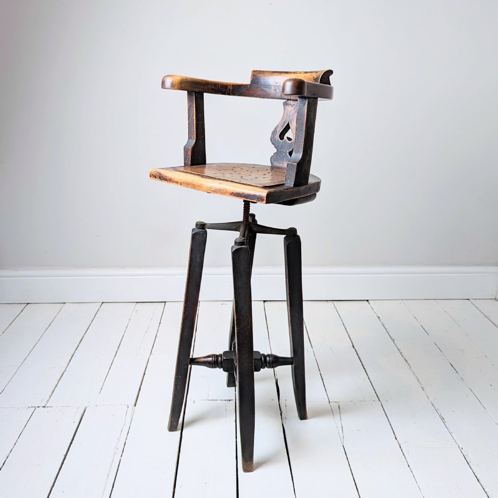 An antique dark wood childs barber chair featuring a swivel seat with a pierced pattern, curved arms, and a cast iron screw adjustment mechanism above splayed turned legs.