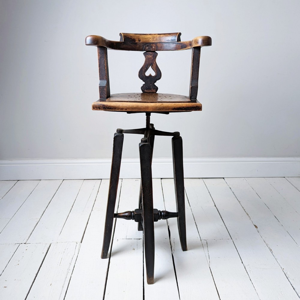 An antique dark wood childs barber chair featuring a swivel seat with a pierced pattern, curved arms, and a cast iron screw adjustment mechanism above splayed turned legs. View II