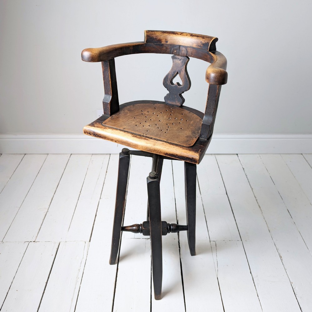 An antique dark wood childs barber chair featuring a swivel seat with a pierced pattern, curved arms, and a cast iron screw adjustment mechanism above splayed turned legs. View V