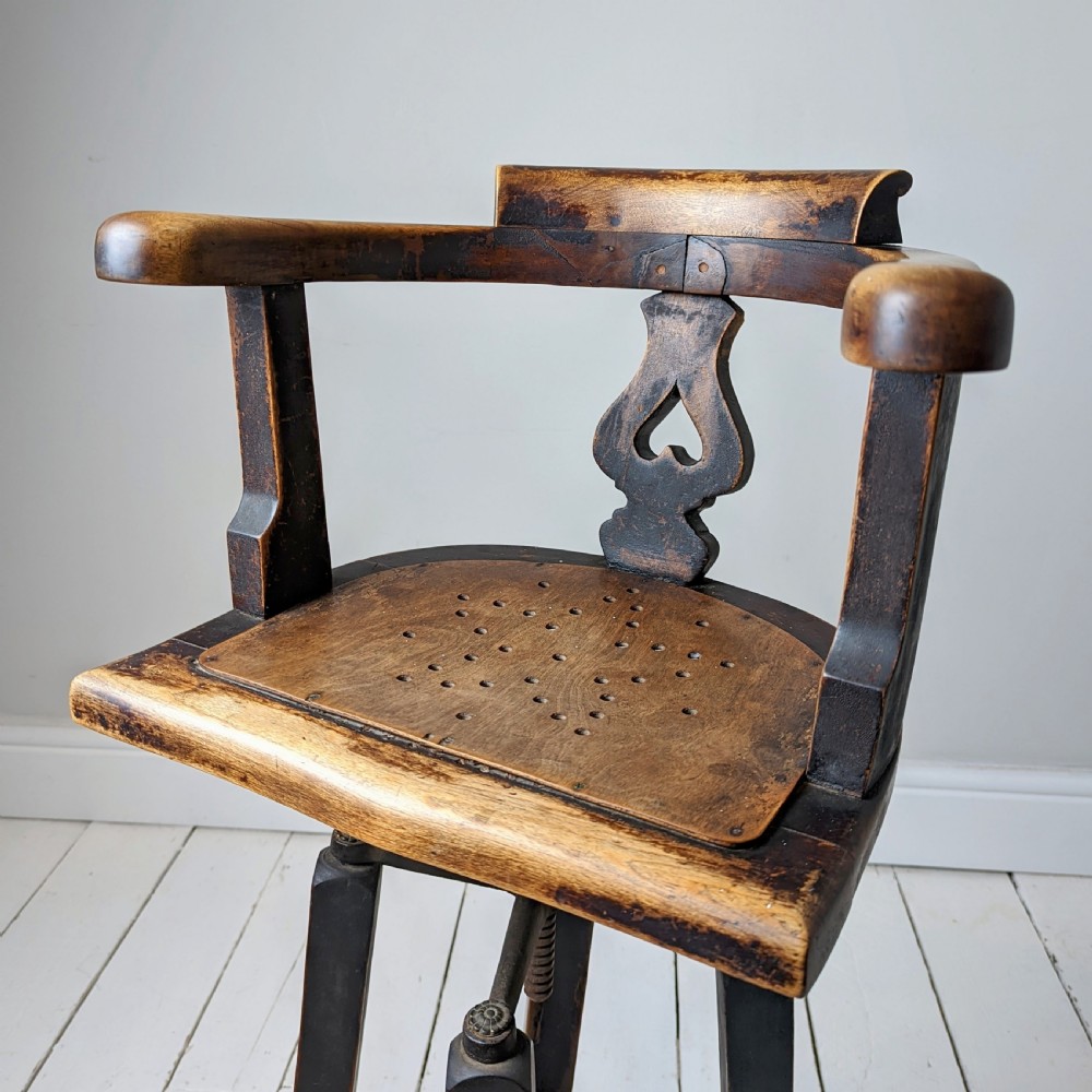 An antique dark wood childs barber chair featuring a swivel seat with a pierced pattern, curved arms, and a cast iron screw adjustment mechanism above splayed turned legs. View VI