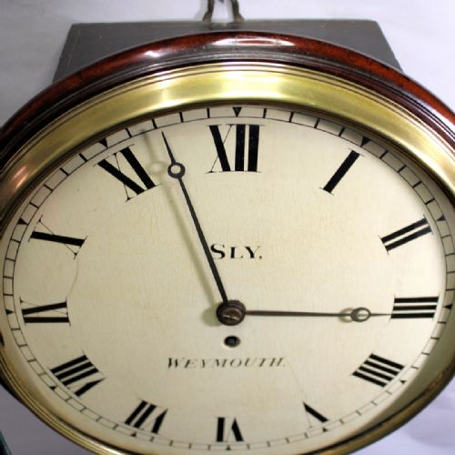 fig 2. A circular antique mahogany wall clock with a brass bezel, off-white dial displaying black Roman numerals, and maker signature Sly Weymouth.