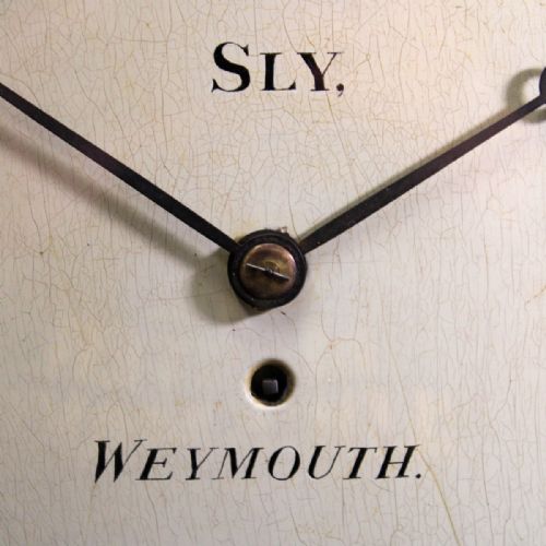 fig 3. A circular antique mahogany wall clock with a brass bezel, off-white dial displaying black Roman numerals, and maker signature Sly Weymouth.
