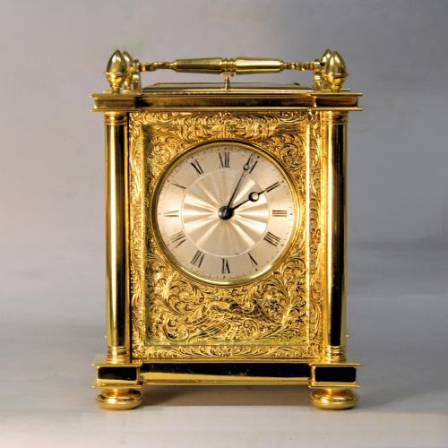 fig 1. An ornate Victorian gilt brass carriage clock featuring detailed foliage engraving, corner columns, a decorative scroll handle, and a silvered sunburst dial with Roman numerals.
