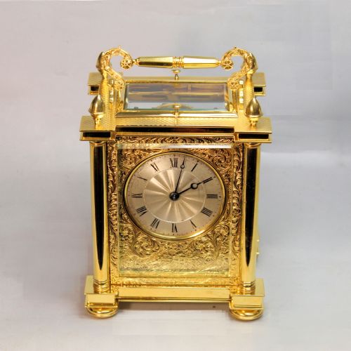 fig 2. An ornate Victorian gilt brass carriage clock featuring detailed foliage engraving, corner columns, a decorative scroll handle, and a silvered sunburst dial with Roman numerals.