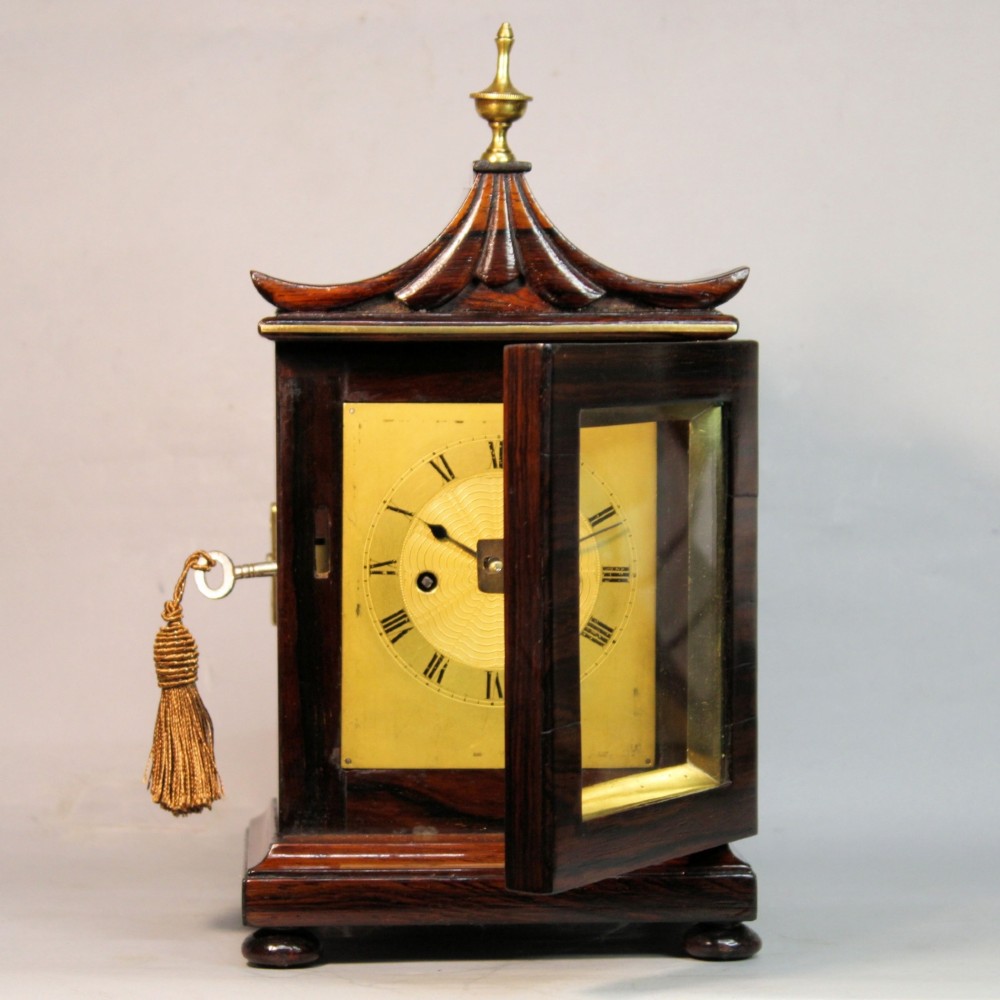 A Victorian rosewood miniature bracket clock featuring a pagoda roof, brass finial, and engine-turned brass dial alongside a matching carved rosewood wall bracket. View IV