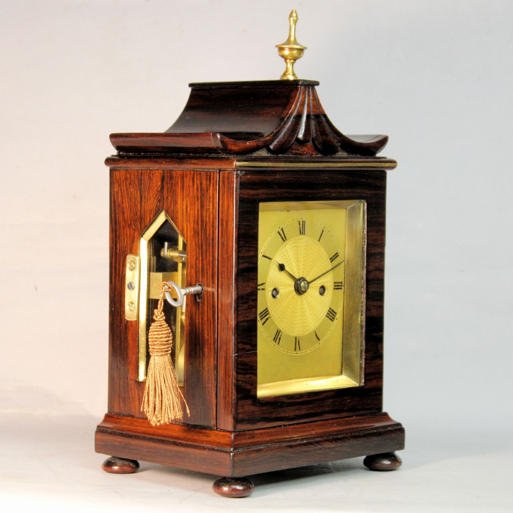 A Victorian rosewood miniature bracket clock featuring a pagoda roof, brass finial, and engine-turned brass dial alongside a matching carved rosewood wall bracket. View V