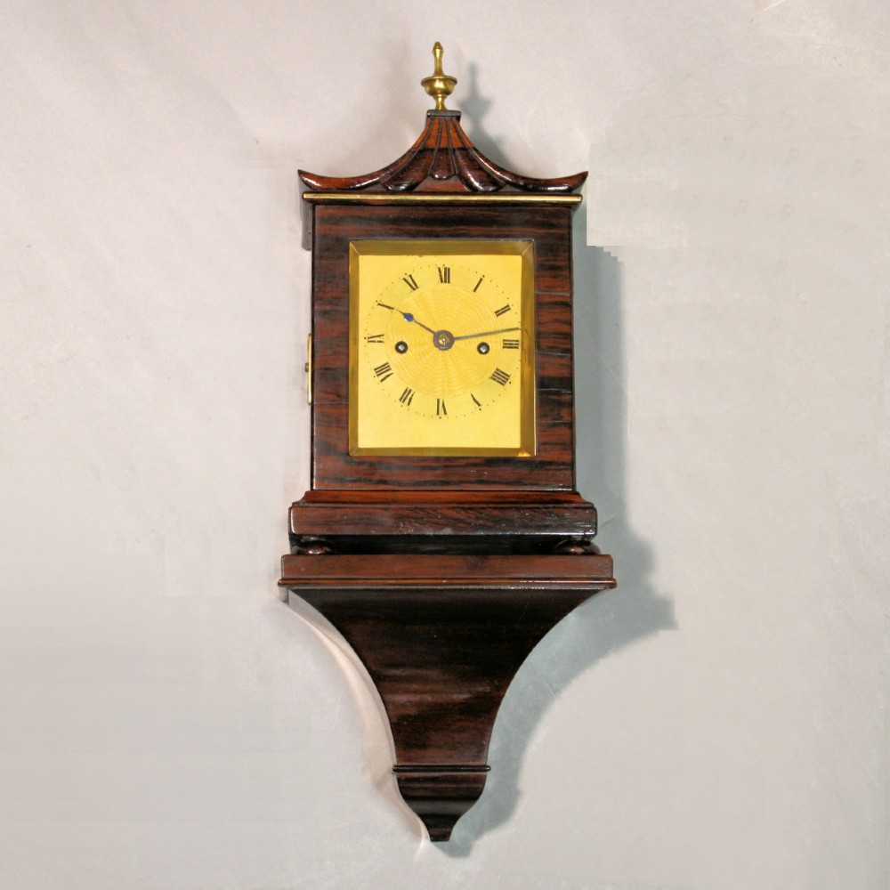 A Victorian rosewood miniature bracket clock featuring a pagoda roof, brass finial, and engine-turned brass dial alongside a matching carved rosewood wall bracket. View VI