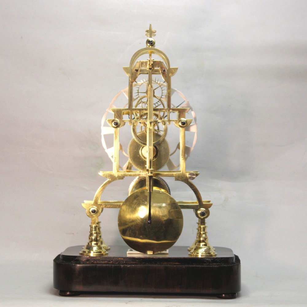 A Victorian brass skeleton clock featuring exposed internal gears, a silvered chapter ring, and a glass dome on a dark wooden base. View VI