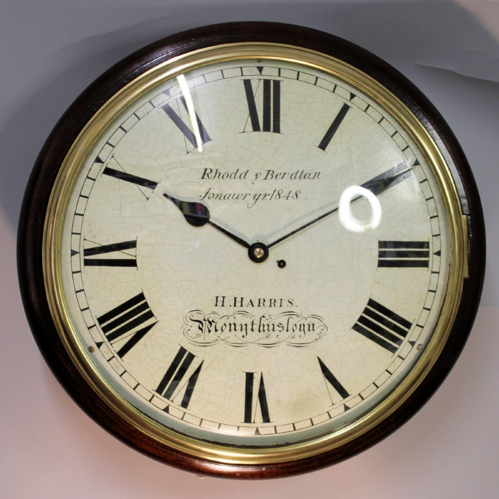 A circular Victorian wall clock featuring a dark mahogany surround, brass bezel, and cream painted dial with Roman numerals and black metal hands. View IV