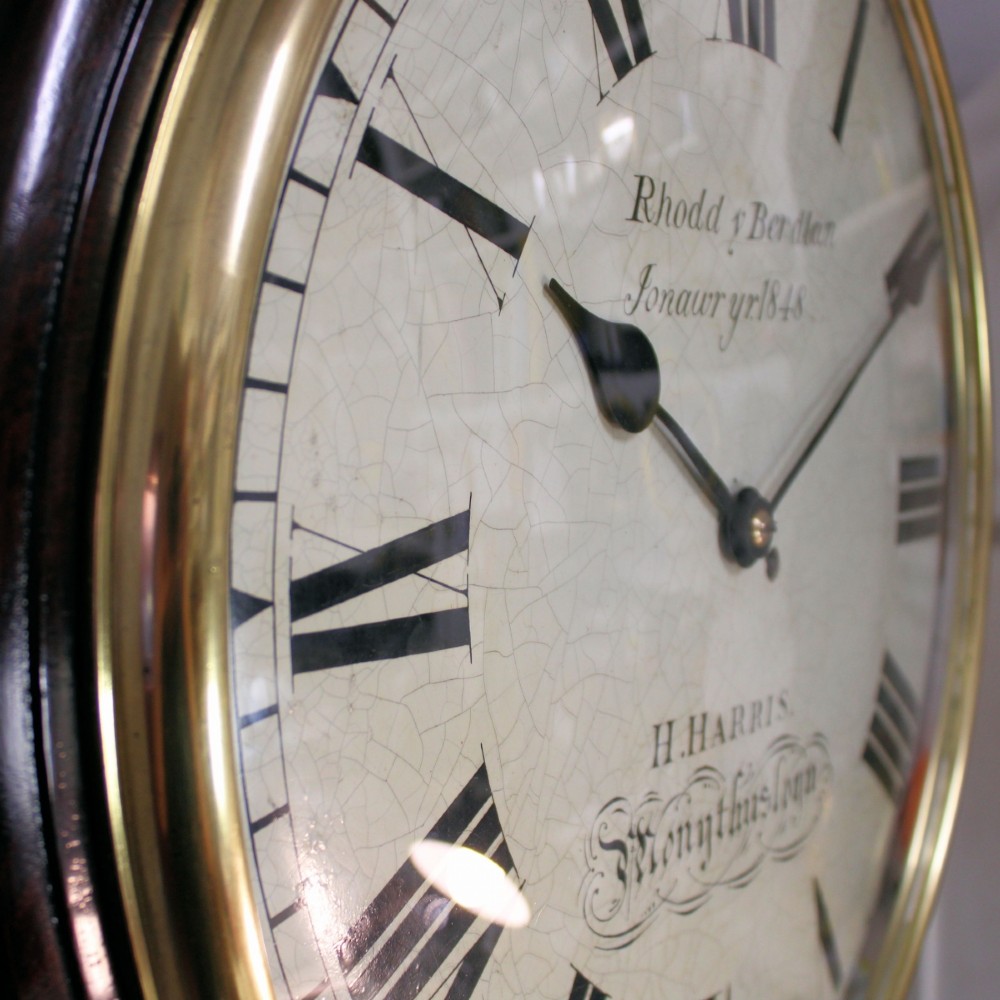 A circular Victorian wall clock featuring a dark mahogany surround, brass bezel, and cream painted dial with Roman numerals and black metal hands. View V
