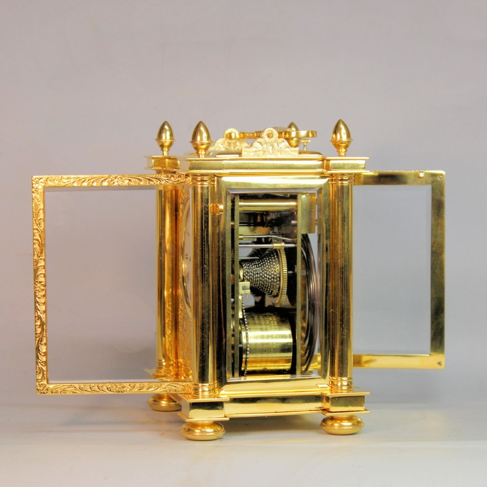 An ornate Victorian gilt brass carriage clock featuring detailed foliage engraving, corner columns, a decorative scroll handle, and a silvered sunburst dial with Roman numerals. View X