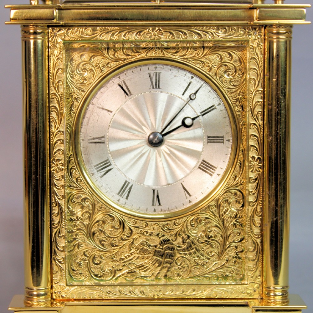 An ornate Victorian gilt brass carriage clock featuring detailed foliage engraving, corner columns, a decorative scroll handle, and a silvered sunburst dial with Roman numerals. View XII
